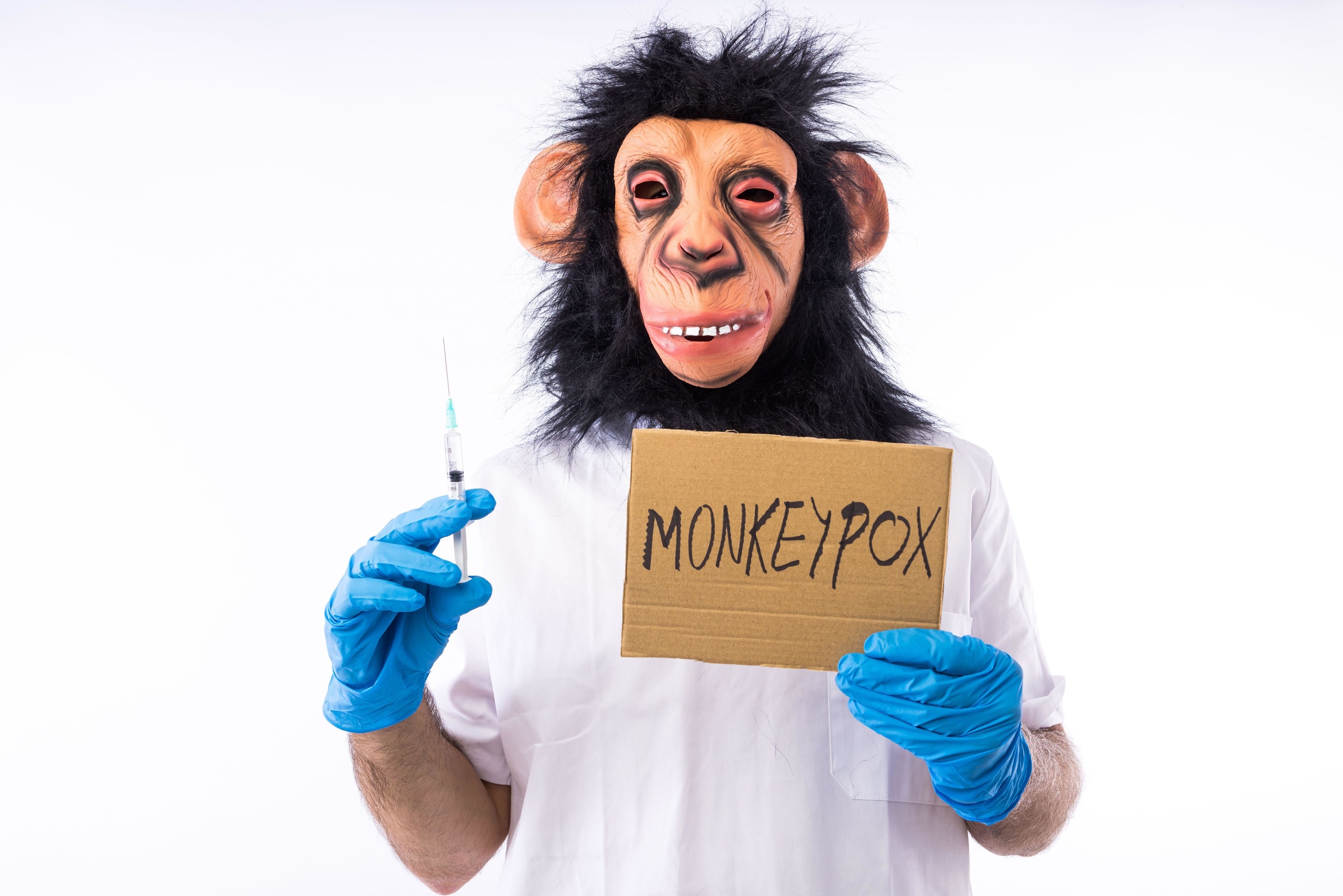 A man wearing a doctor&#x27;s coat and gloves, holding a syringe and a monkeypox sign while wearing a monkey mask
