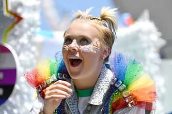JoJo Siwa wears a jean jacket with rainbow-colored tule on the shoulders and matching face gems with her hair in ponytails.