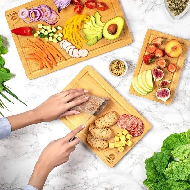 Person cutting a baguette and charcuterie and chopped fruits and veggies on cutting boards