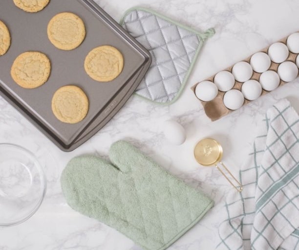 Mint colored oven mit and pot holder next to baked cookies, eggs and baking items