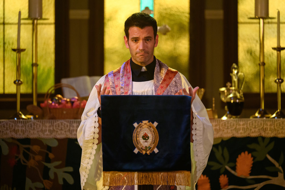 Colin Donnell stands by an altar dressed as a priest
