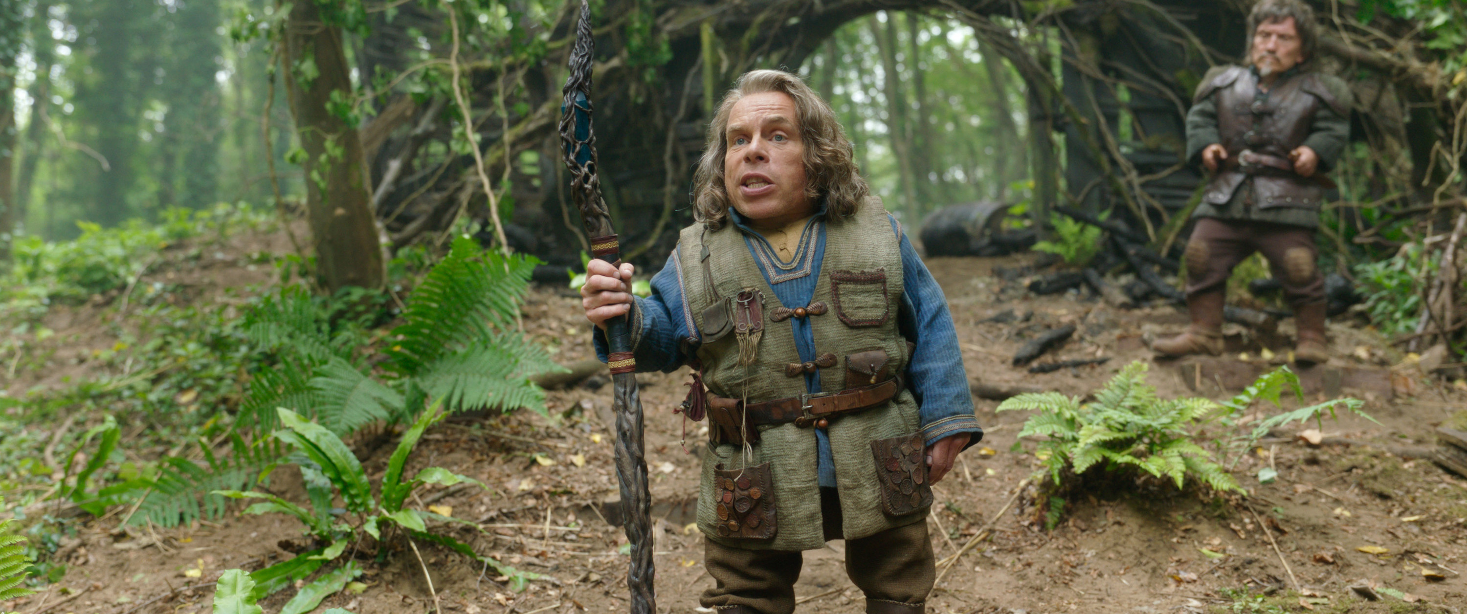 Warwick Davis stands in the woods with a walking stick
