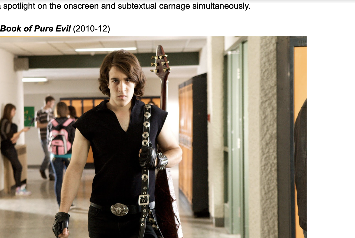 A brown-haired teenager in all-black clothes and a heavy metal guitar walks down a high school hallway