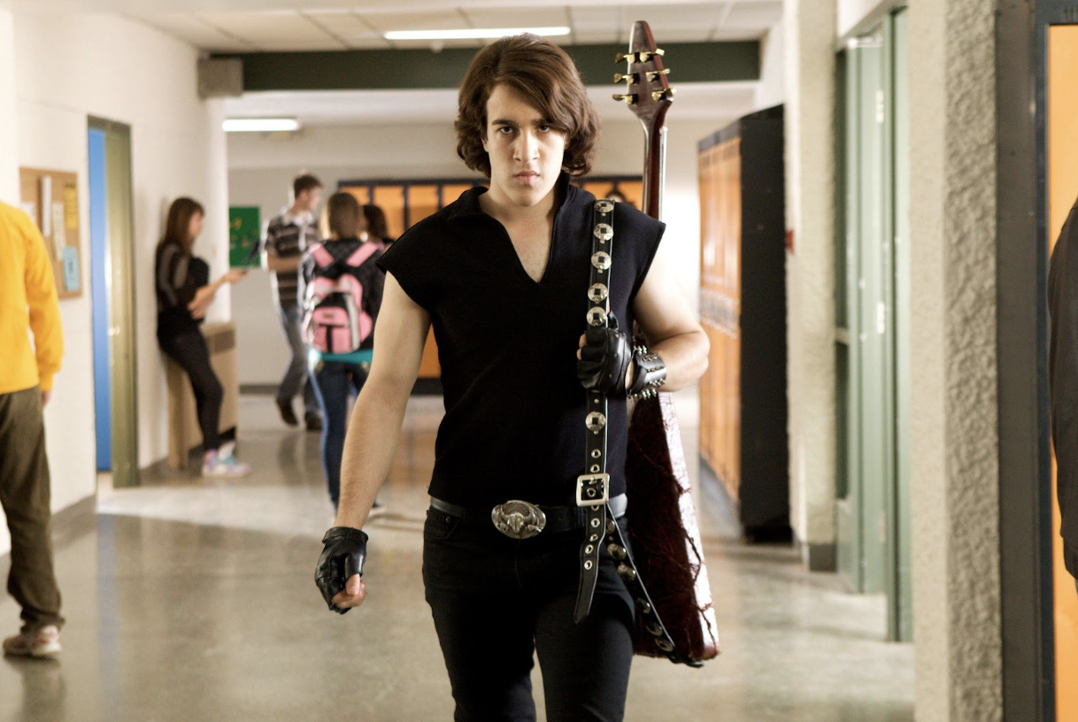 A brown-haired teenager in all-black clothes and a heavy metal guitar walks down a high school hallway
