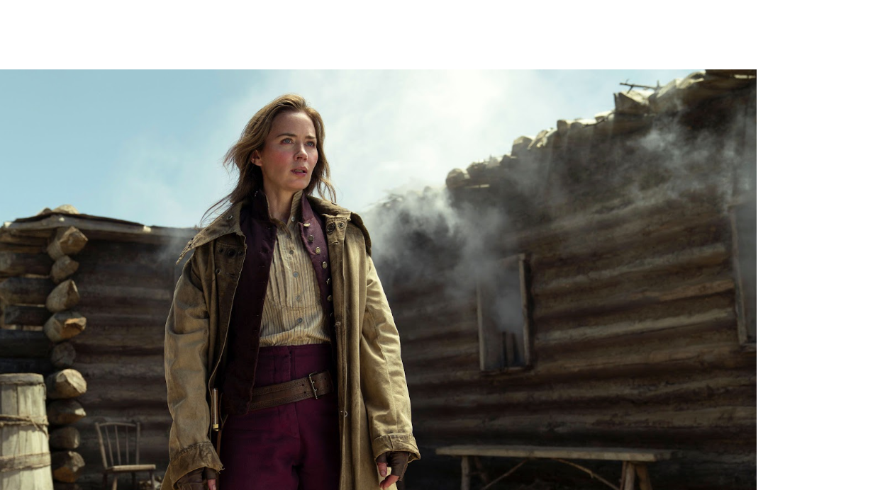 A woman in old western clothes stands stoically outside of a smoky cabin.