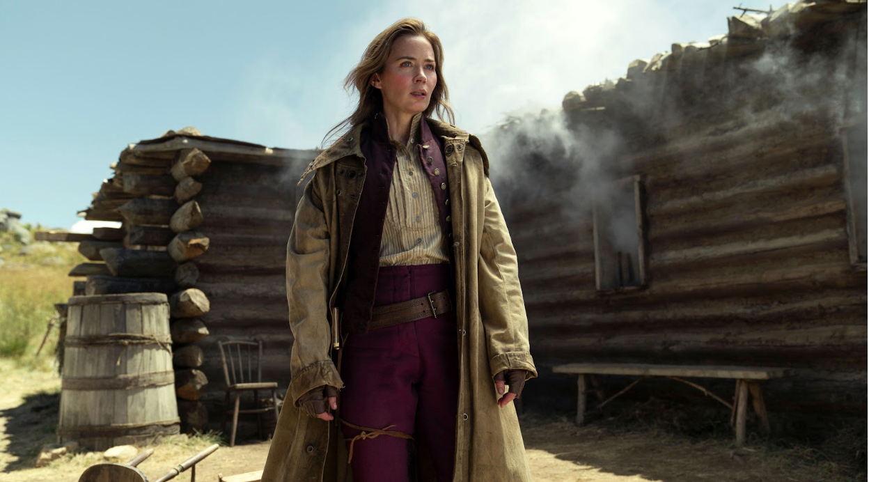 A woman in old western clothes stands stoically outside of a smoky cabin.
