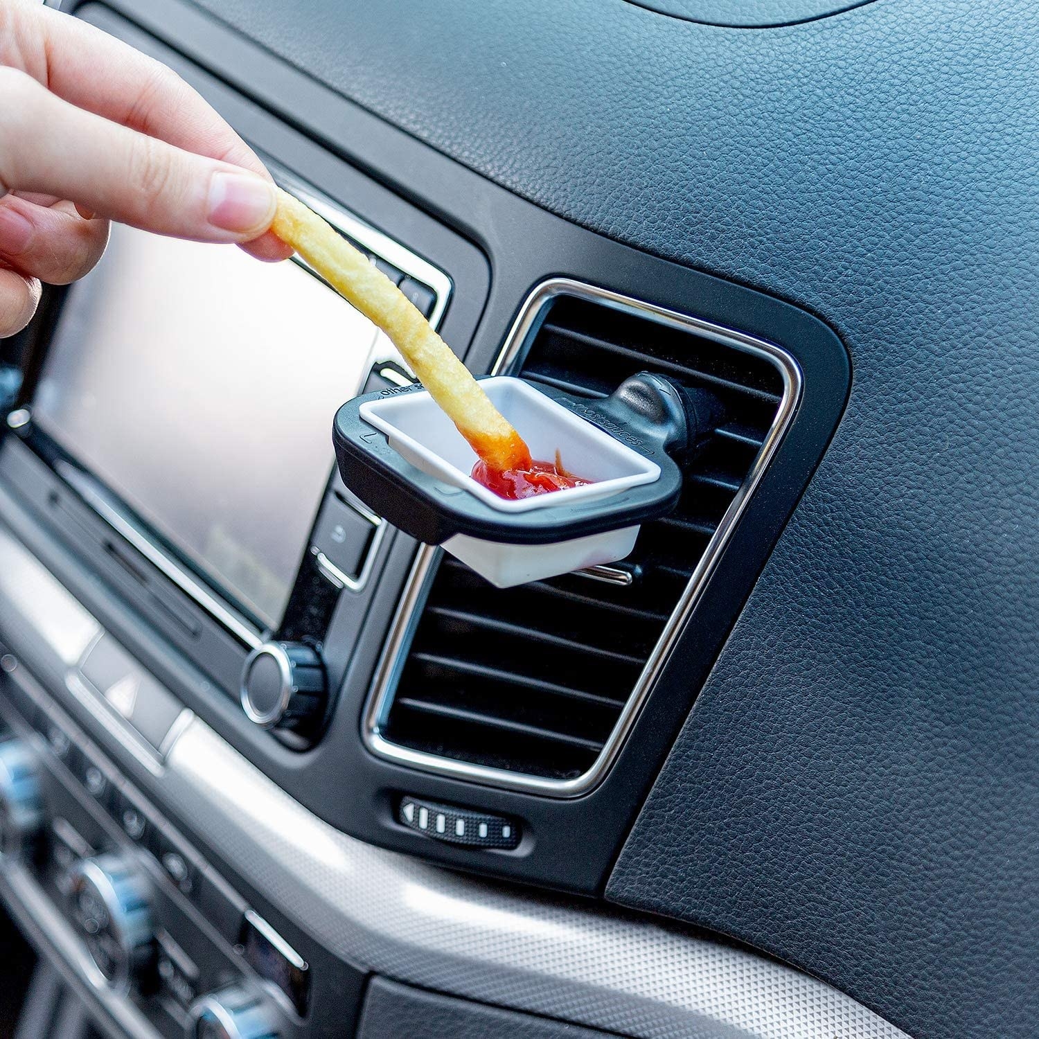 the sauce holder clipped to a car vent with ketchup in it and someone dipping a fry into it