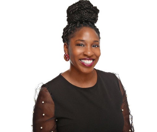 A photo of Raquel Martin, Ph.D., HSP. She is a Black woman, smiling warmly at the camera. She has her twists in a top knot. She is wearing a black shirt with sheer sleeves decorated in pearls. She has round earrings with a pattern.