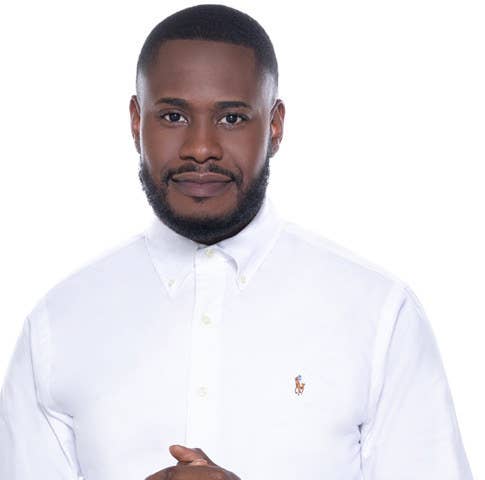 A photo of Arron Muller, a Black man looking kindly at the camera. He has a small smile, a shaved hear with a fade, and a well-kept beard. He is in a white button down and his hands are clasped