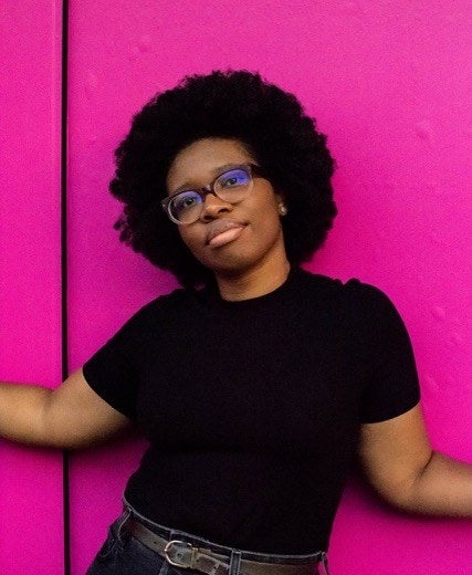 Grace Johnson, a Black woman posing in front of a neon pink background. She has an afro and is wearing all black. She looks relaxed and content