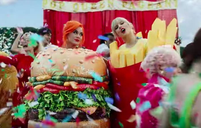 Katy Perry in a burger costume and Taylor Swift in a fries costume stand together while surrounded by people partying