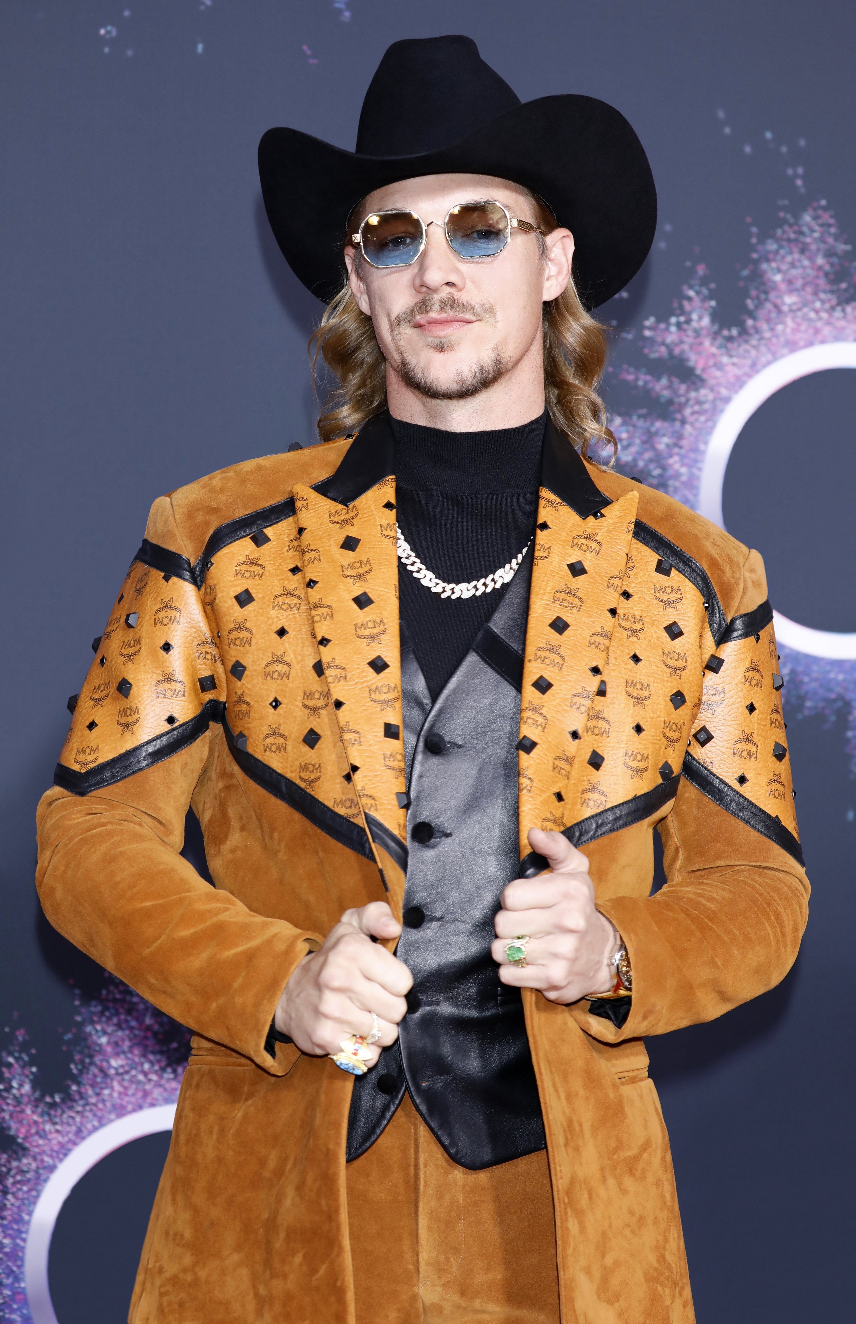 Diplo poses on the red carpet in a tan suede suit, with a black cowboy hat and blue sunglasses