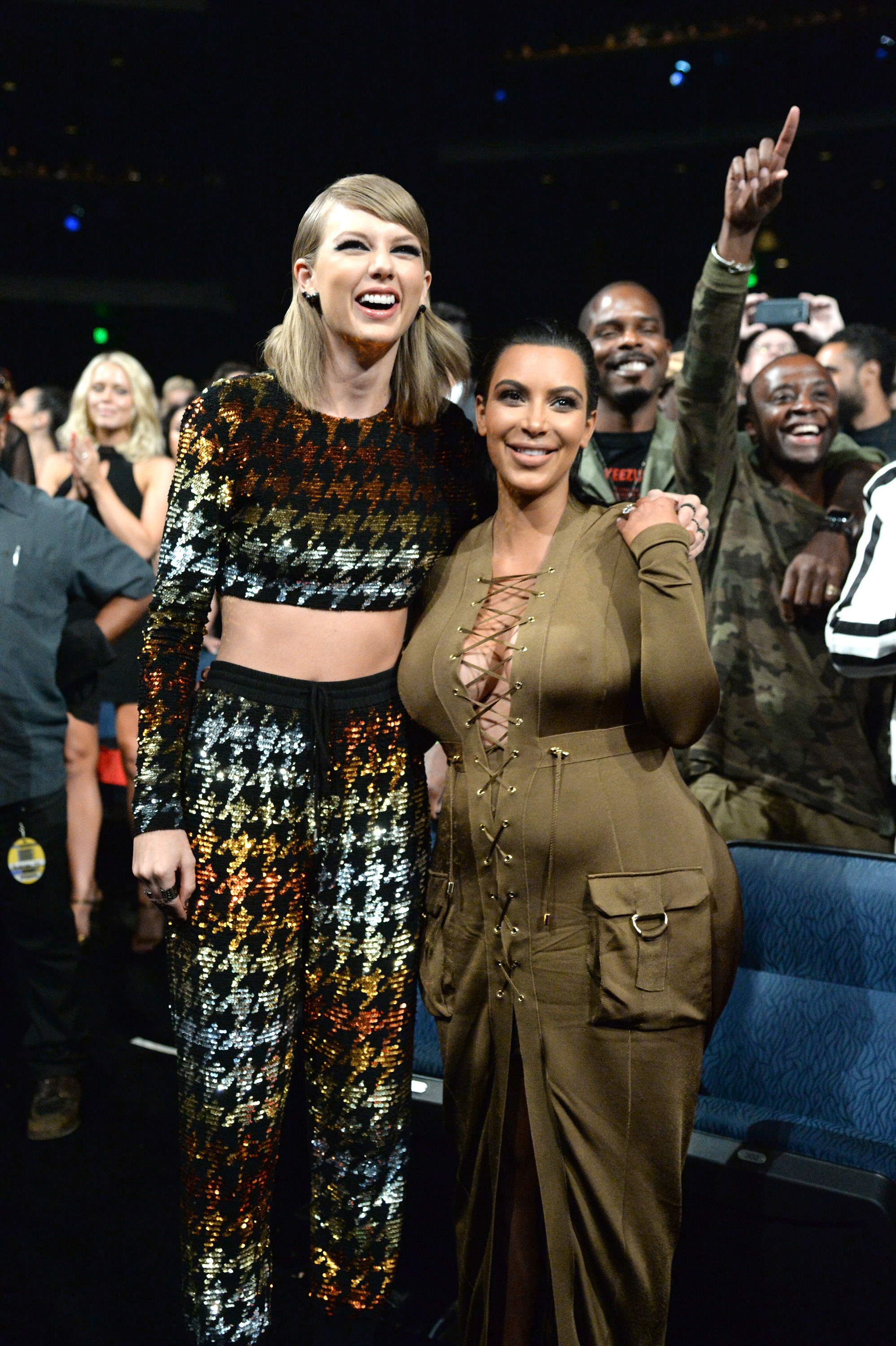 Taylor Swift in a sparkly jumpsuit and Kim Kardashian in a khaki dress stand front row at an awards show, smiling with their hands around each other