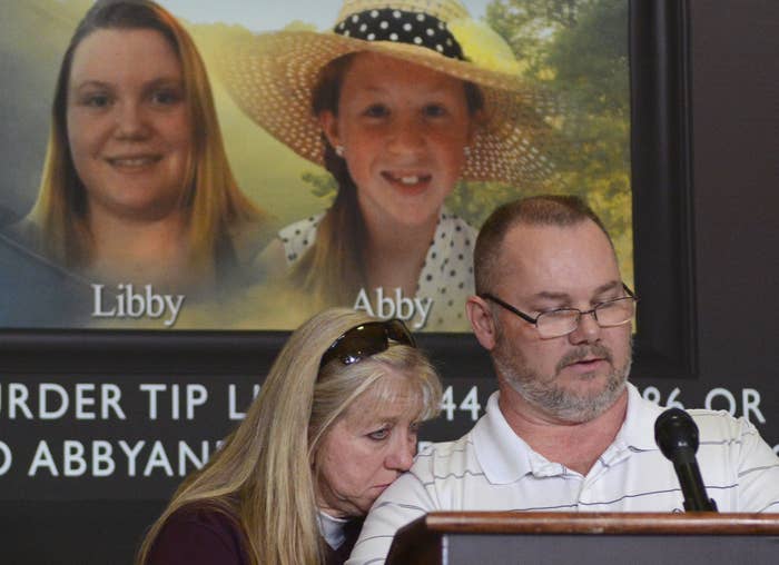 A man stands behind a lectern with a microphone and speaks as a woman wearing sunglasses on her head presses her face into his shoulder. They are both standing in front of an image of Libby and Abby, two young girls, one wearing a straw hat