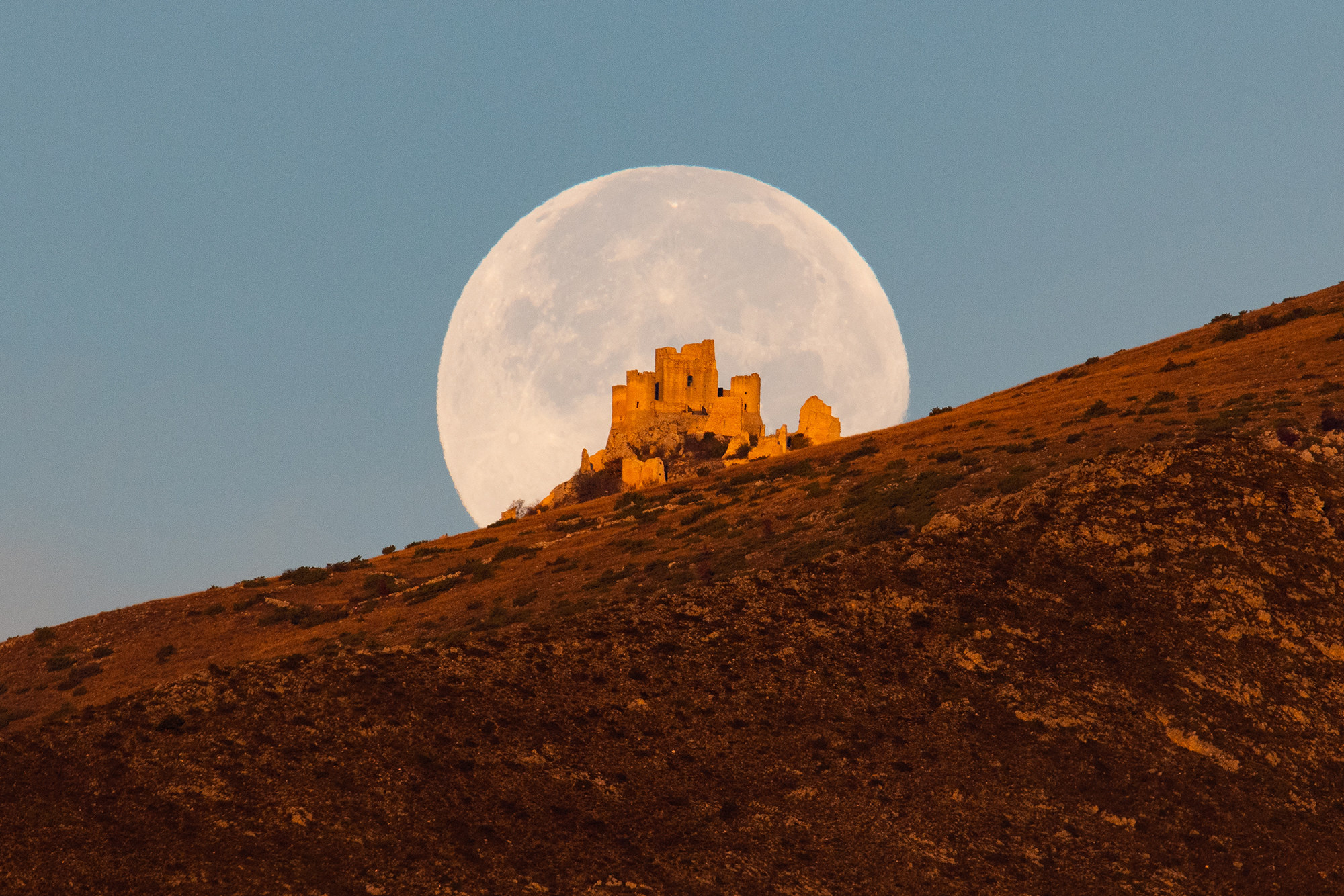 A full white moon sits behind a castle on a hill