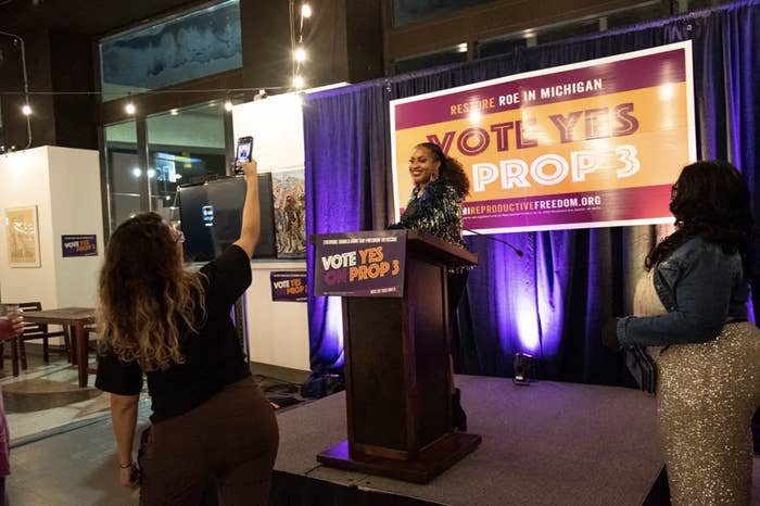 A woman at a podium stands in front of a sign that reads "vote yes on prop 3" as someone takes her photo