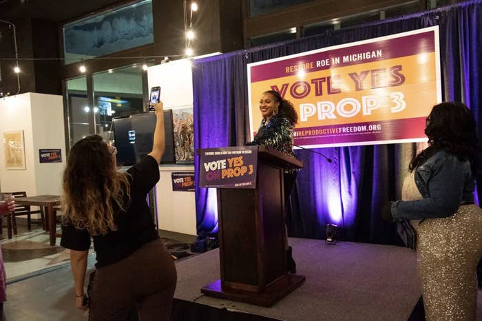 A woman at a podium stands in front of a sign that reads &quot;vote yes on prop 3&quot; as someone takes her photo