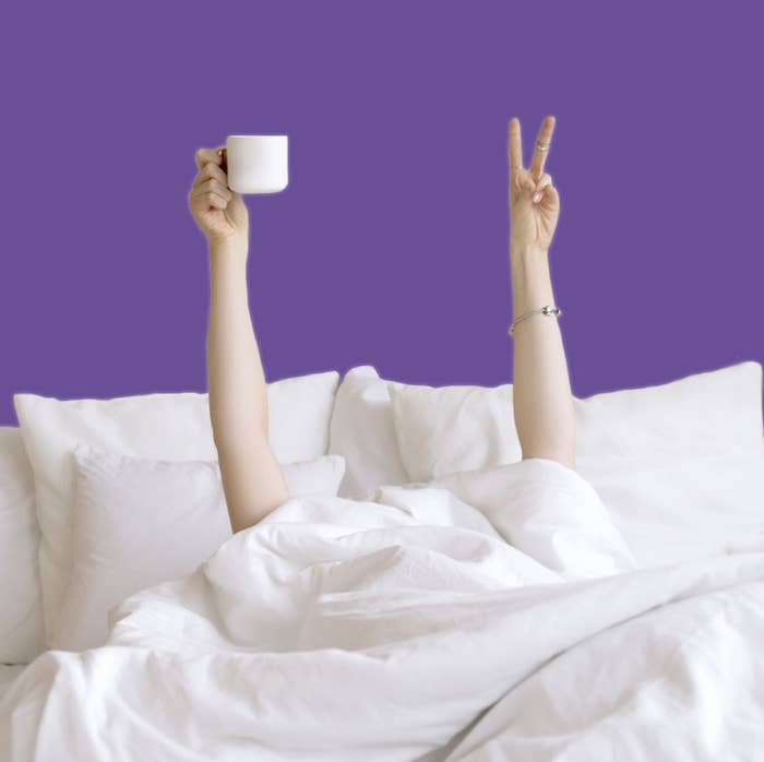 a person hiding under their duvet in bed with a cup of coffee in their hand