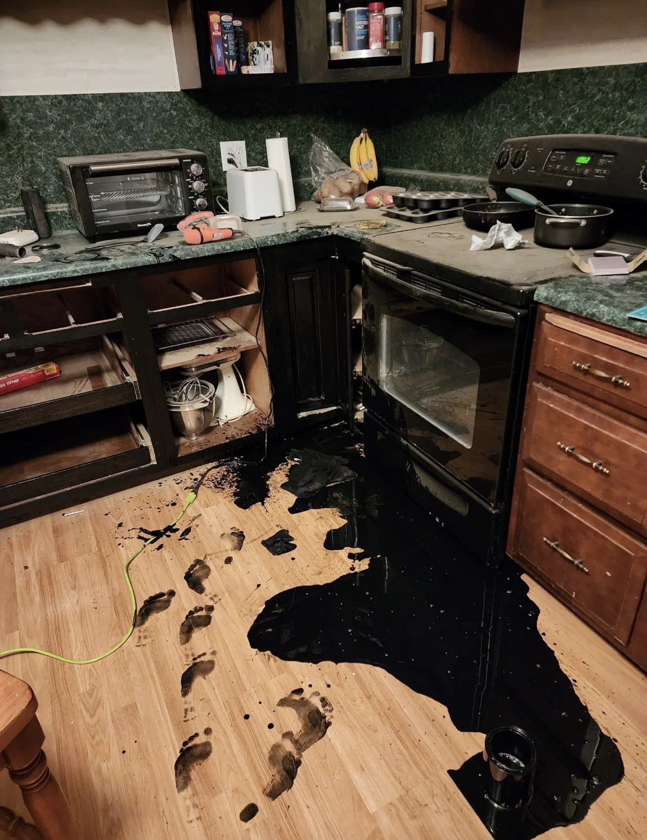 A kitchen with a thick liquid leaking out of the bottom of the oven, and footprints all over the floor that match the color of the liquid