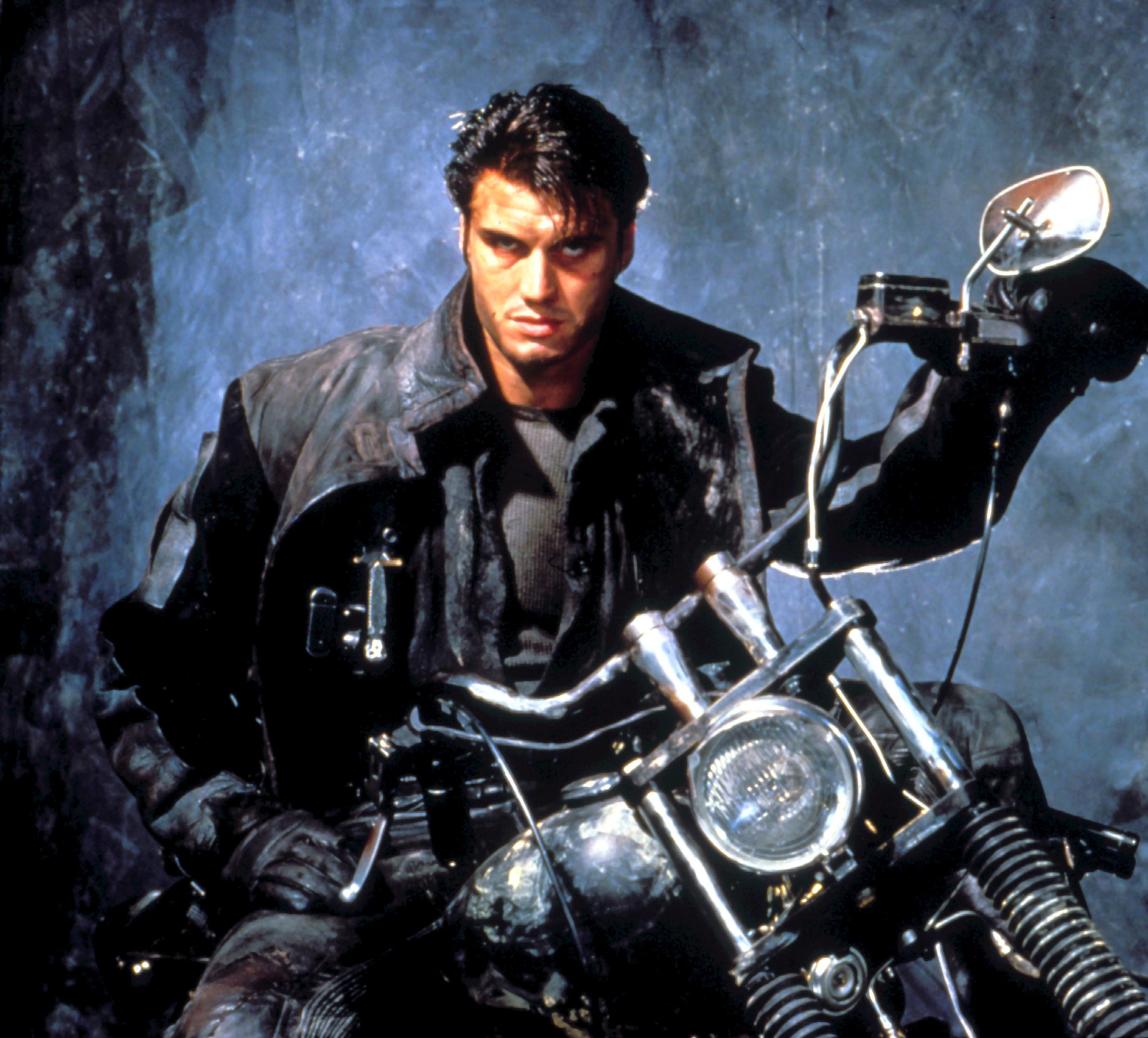 An imposing man in tattered leathers sits atop a motorcycle with a haunted stare