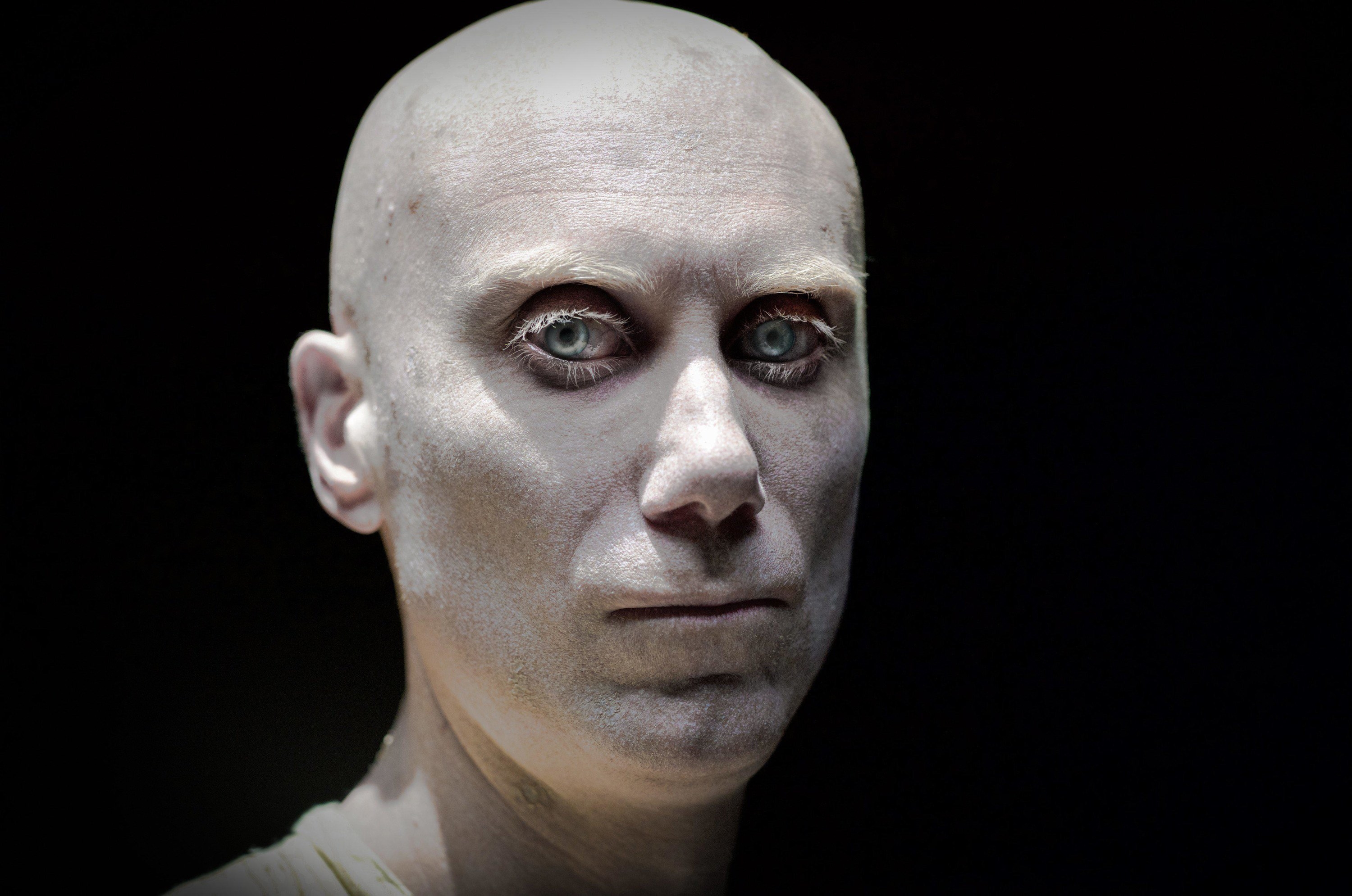 A bald albino mutant stares directly into the camera against a black background