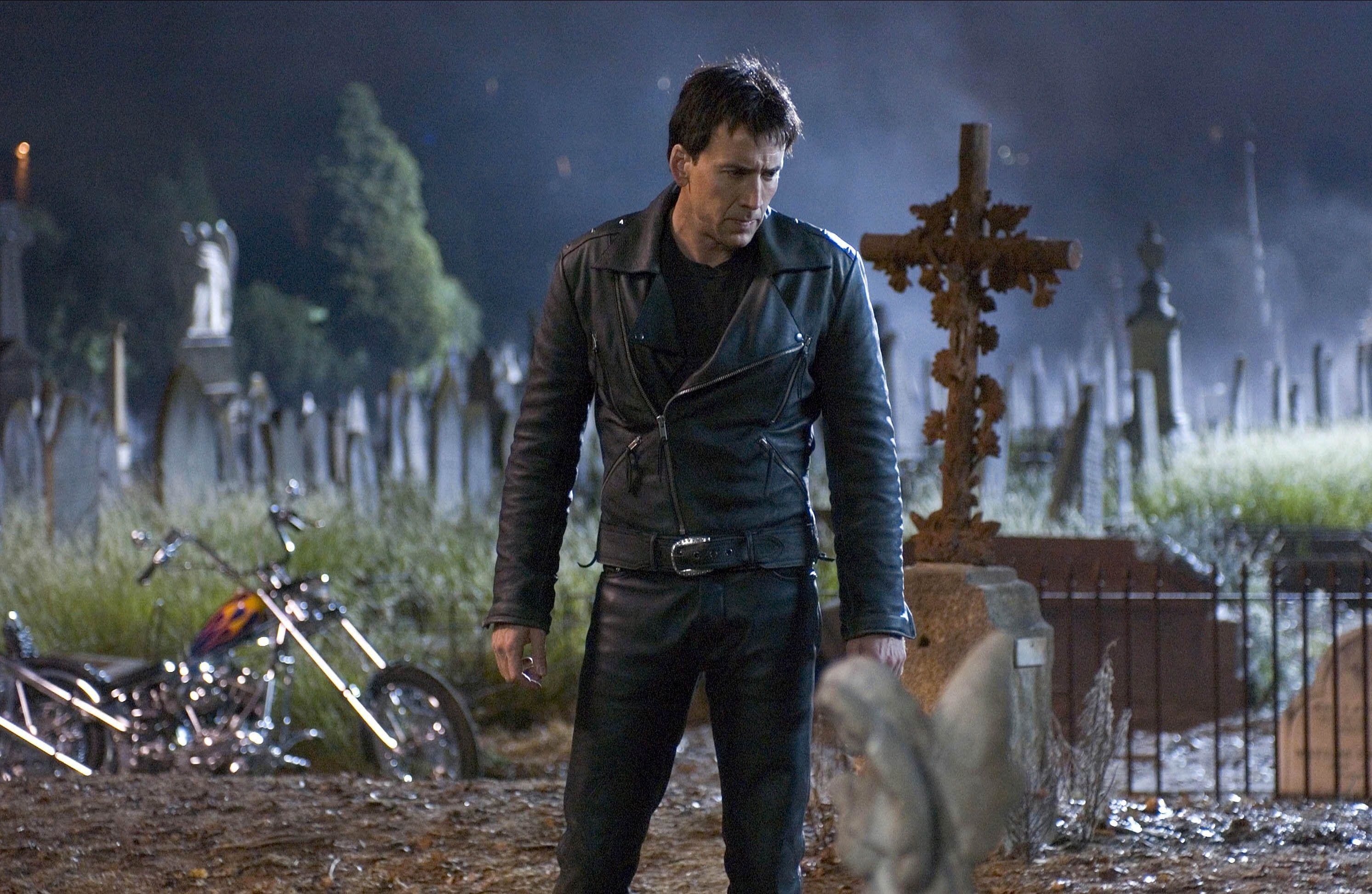 A stoic man in leathers stands nearby a motorcycle at a graveyard