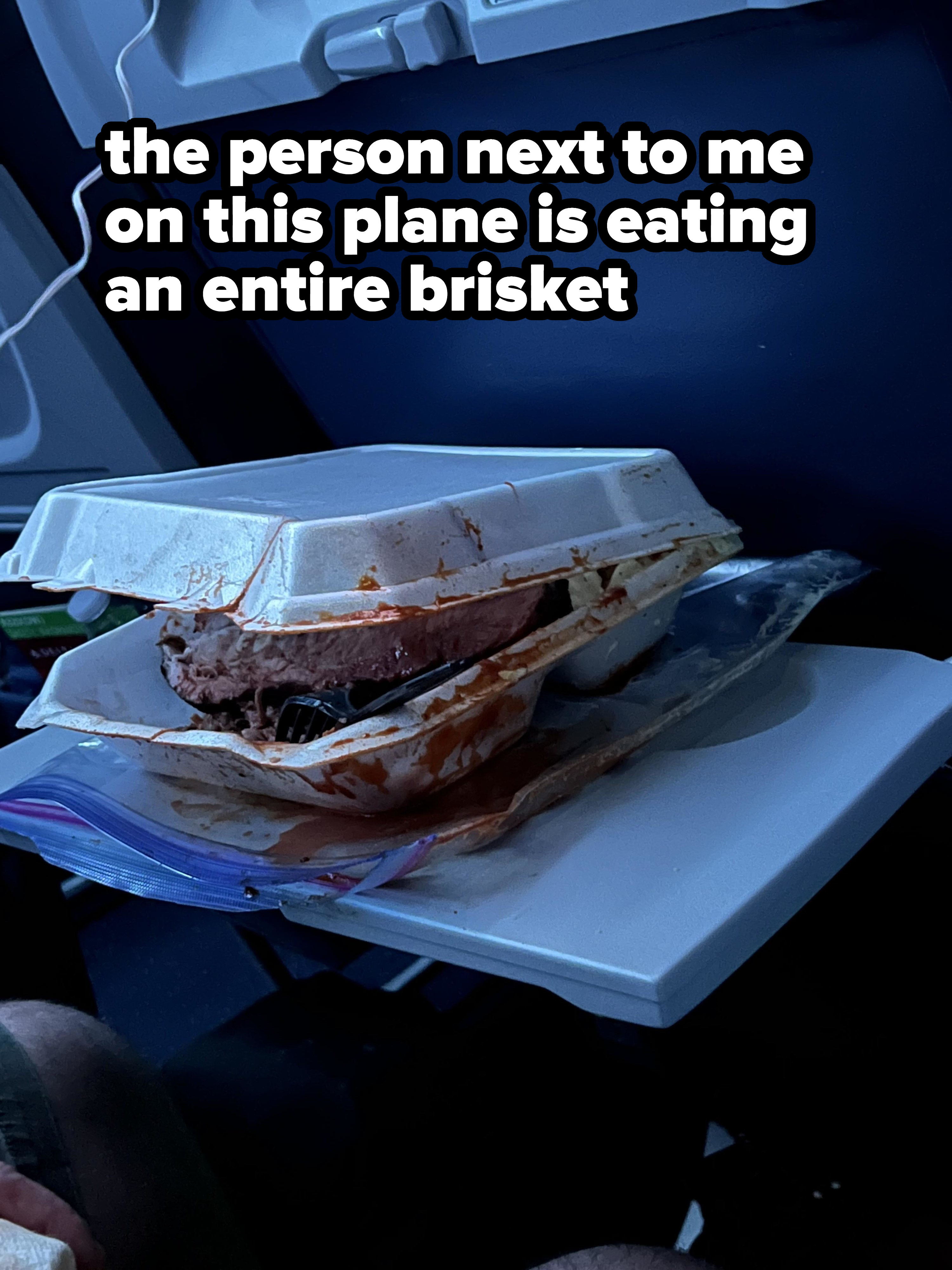 A Styrofoam container with a large piece of meat and gravy/blood extending over the sides and onto the plane&#x27;s seat tray