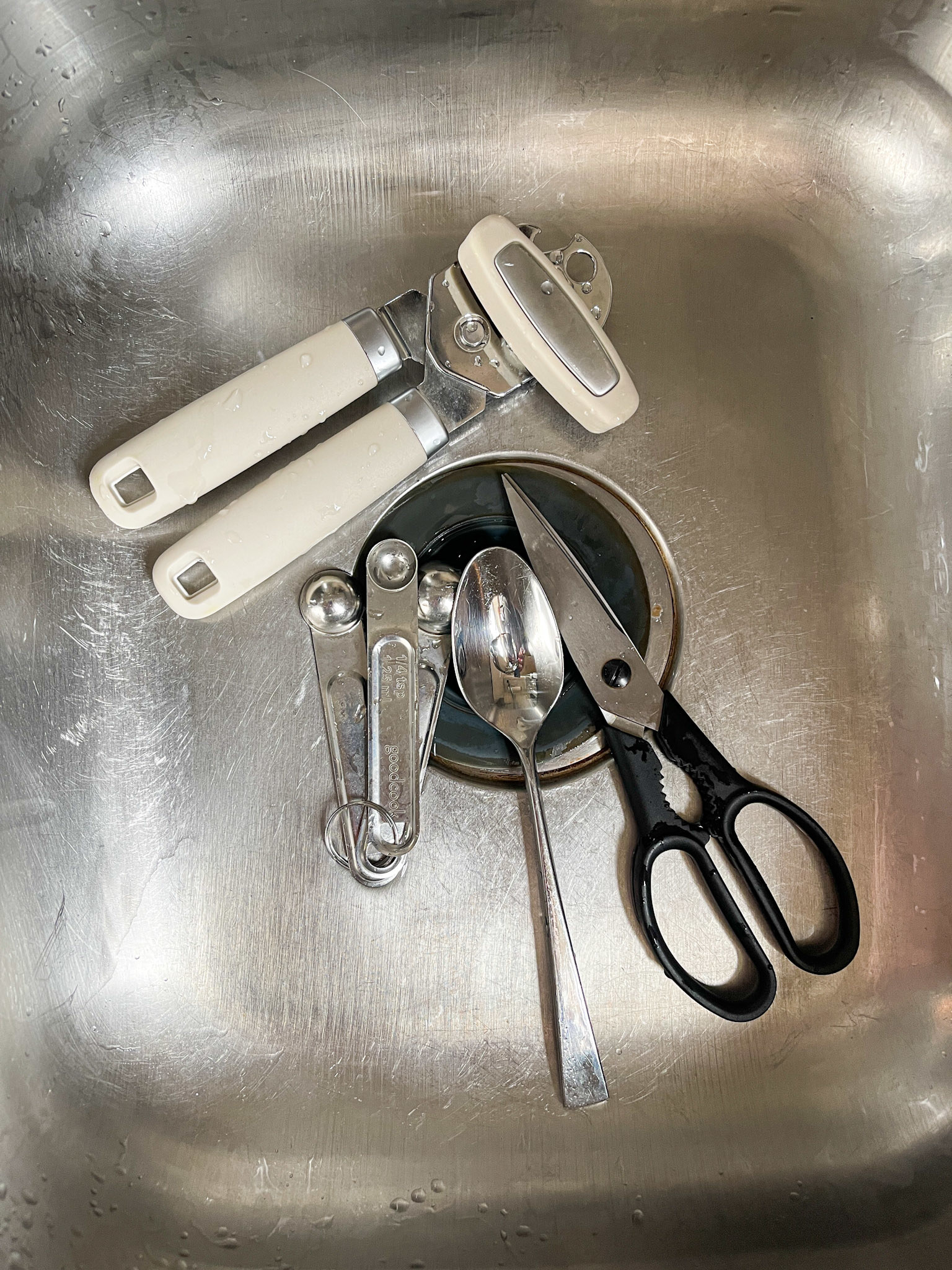 can opener, measuring spoons, teaspoon, and scissors sitting in a steel sink