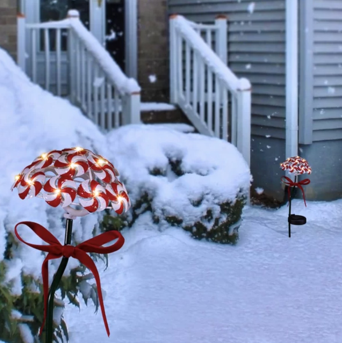 There are two peppermint dome-shaped garden stakes that are lit up with red ribbons placed in the ground near a snowy porch
