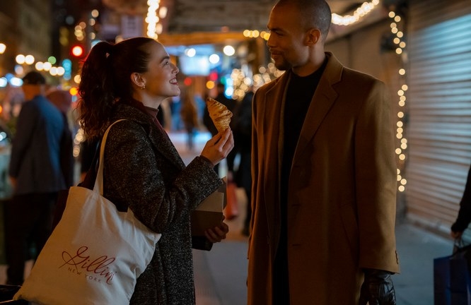 Zoey Deutch and Kendrick Sampson stand opposite each other and smile, Zoey holds a croissant