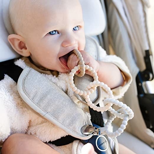 a baby model chewing on the toy, which is clipped to their carseat strap