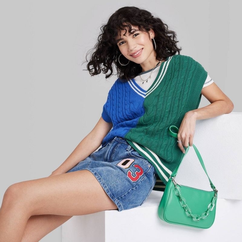 model wearing blue and green sweater vest over white t-shirt with denim mini skirt, holding green purse
