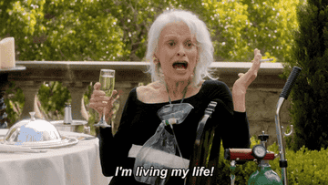A woman holding a cigarette and a glass of champagne and saying, &quot;I&#x27;m living my life!&quot;