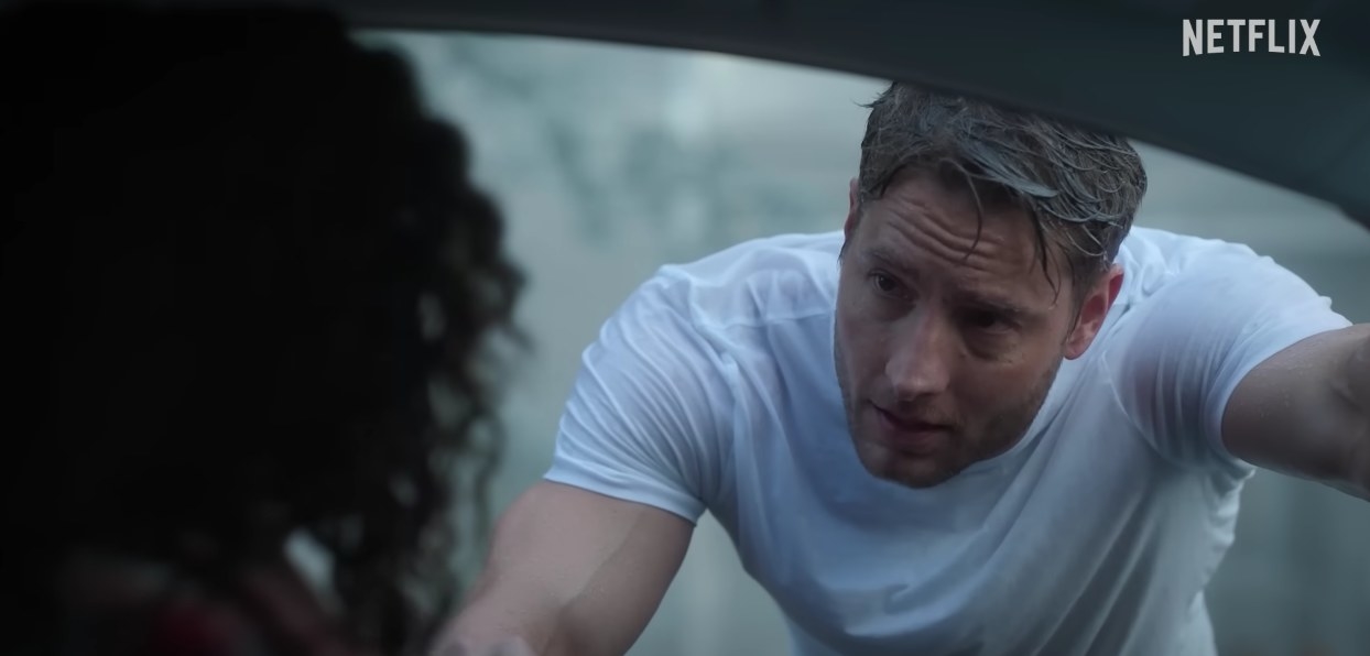 A wet Justin Hartley leans forward and looks into a car window at a woman looking out at him