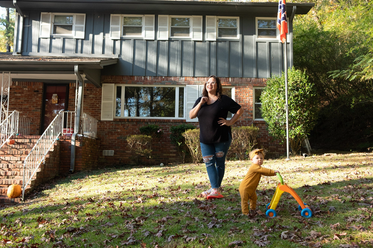 a woman in a black t-shirt and jeans smiles next to her toddler on the front lawn of her house