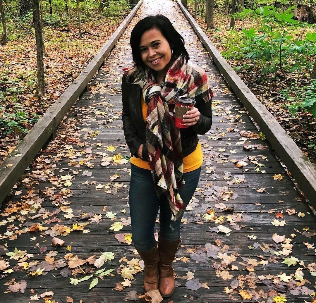 Reviewer standing on fall wooden trail path wearing the red and brown plaid scarf tied over jacket and holding coffee in hand