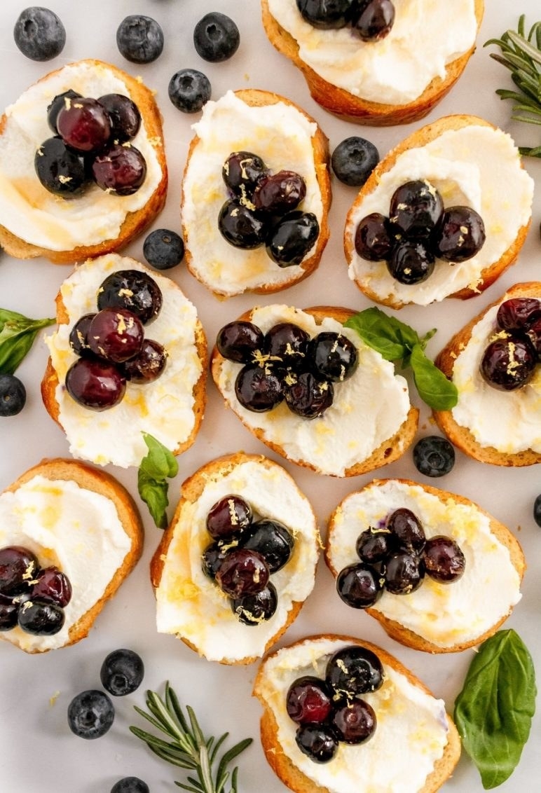 Several blueberry ricotta bruschetta are surrounded by herb sprigs and scattered blueberries