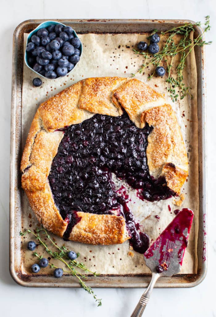 A blueberry galette sits on a baking tray with one slice removed by a serving knife
