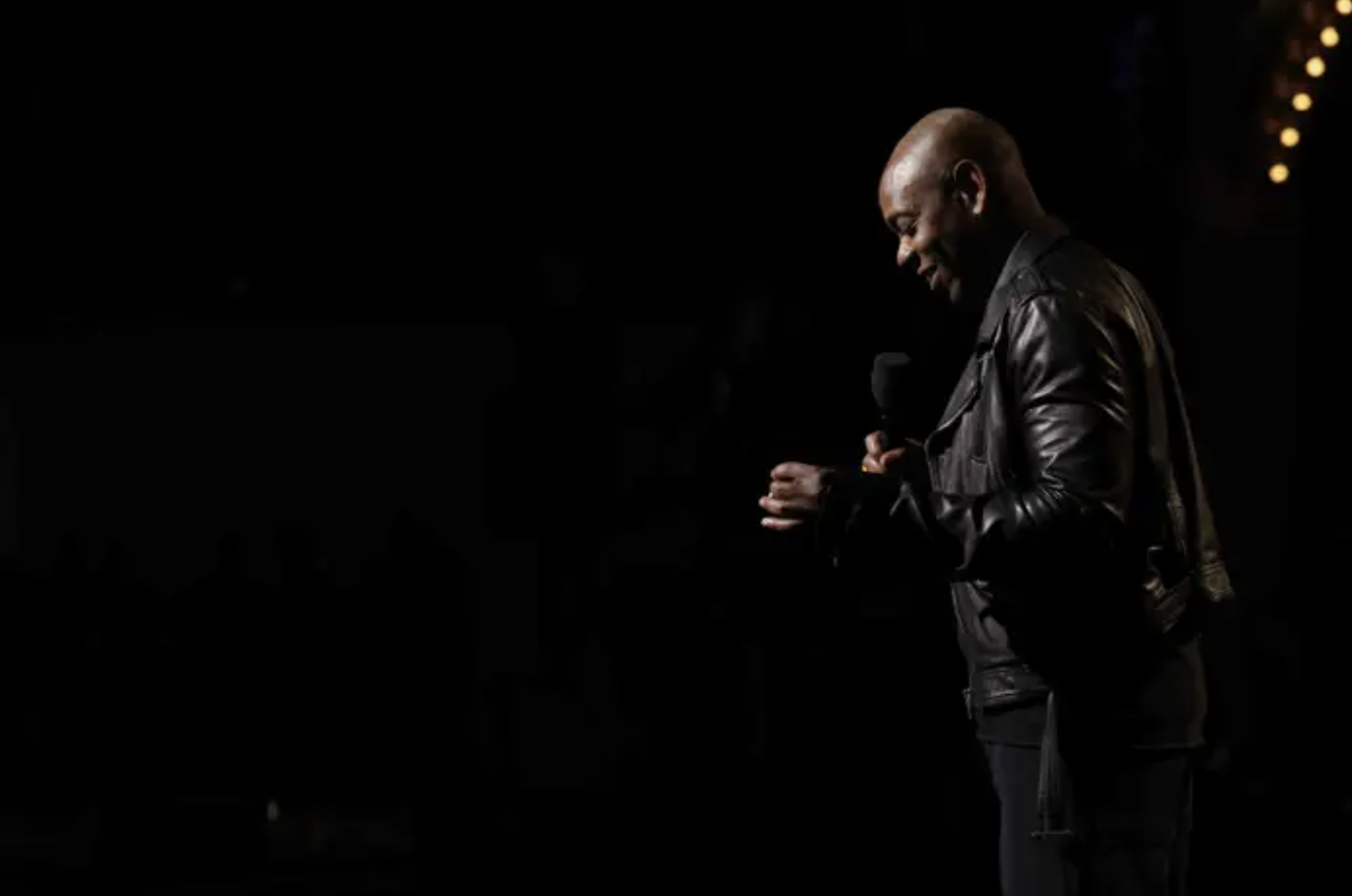 dave chappelle stands in a black leather jacket holding a mic against a pitch black background