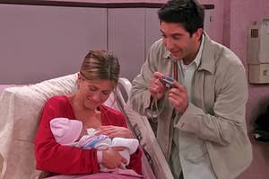 Rachel from Friends holding baby Emma in the hospital bed while Ross stands over them, taking a picture