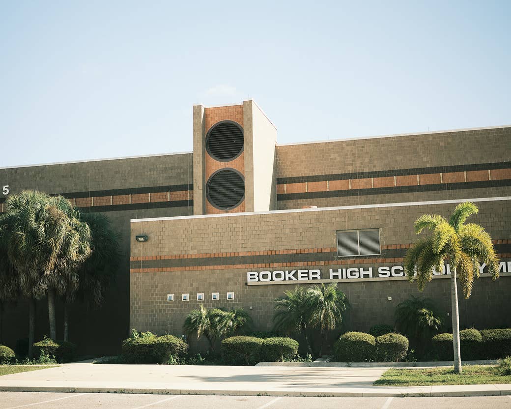A brown, brick school with lighter brown accents. A sign on the school reads &quot;BOOKER HIGH SCHOOL&quot; in a wide sans-serif font. There are palm trees around the school. The building is very dimensional.