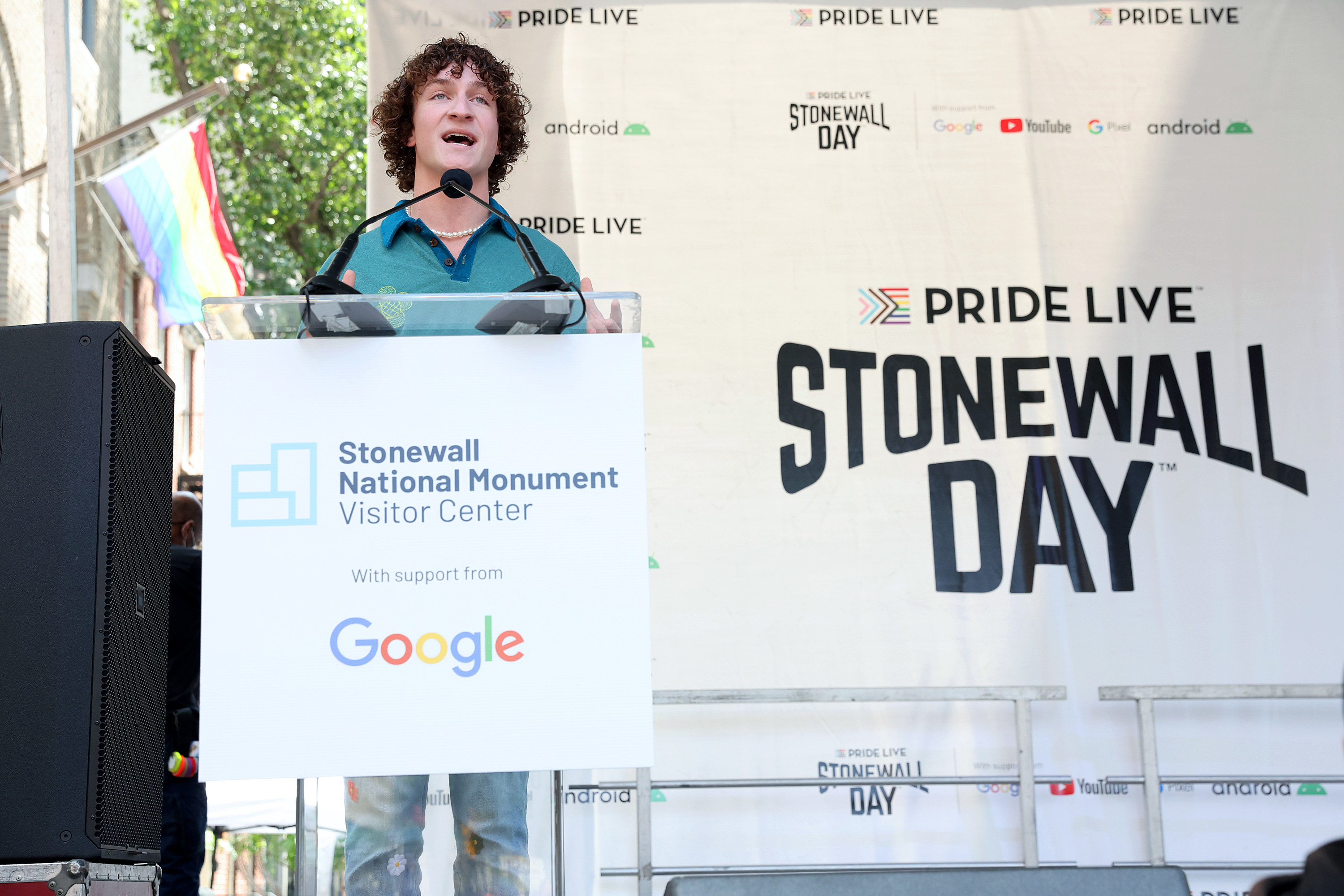 Zander Moricz, a young man with curly brown hair that is speaking at a podium on a stage. The podium says &quot;Stonewall National Monument&quot; and &quot;Google.&quot; the stage says &quot;Stonewall Day&quot;