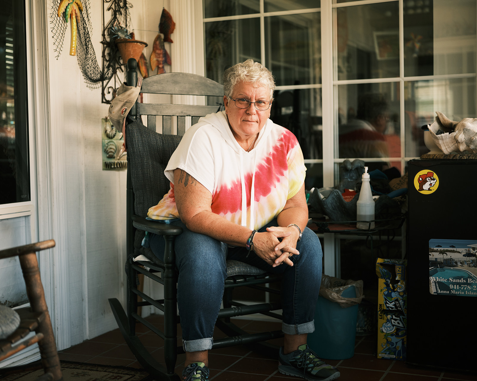 Gail Foreman sits on a rocking chair in a room with bright, natural light. She is leaning forward with her hands clasped in front and her elbows on her knees. 