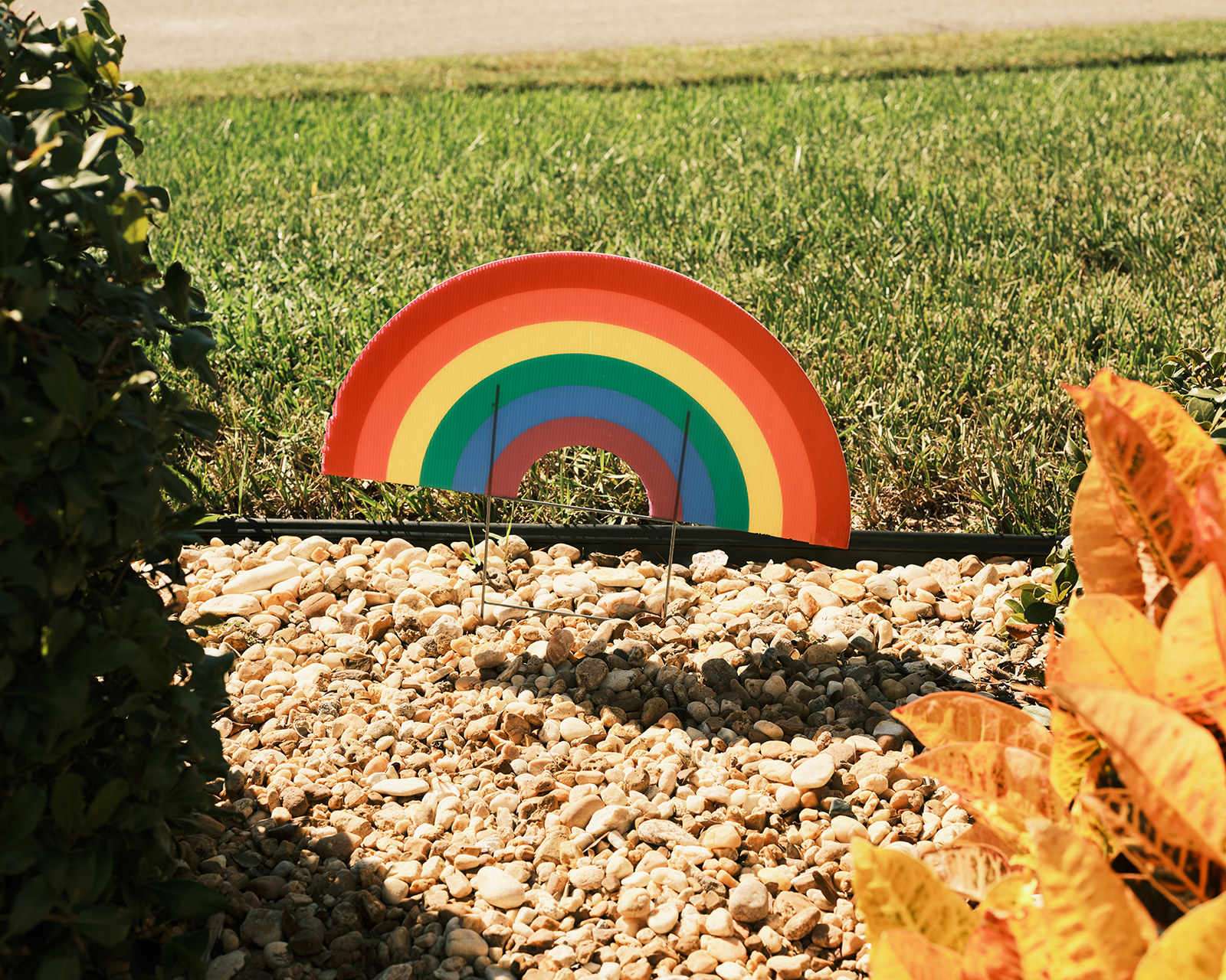 a carboard rainbow in the garden bed outside of Gail Foreman&#x27;s house. There is green grass and the garden bed is comprised of stones.