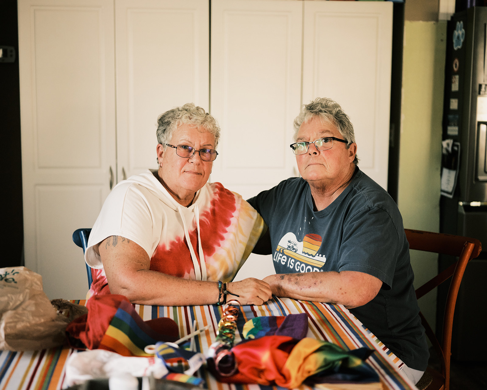 Gail Foreman and her wife, Pat Cummins, sitting at their kitchen table together. Cummins has her arm around Foreman. They both have short gray hair. Foreman has black wire-rim glasses and Cummins has rectangular dark frames.