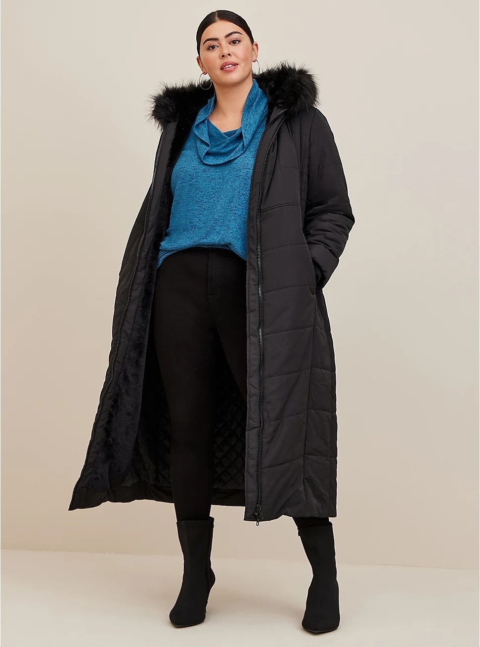 Model wearing long black puffer jacket with fur hood, wearing blue top and black pants underneath