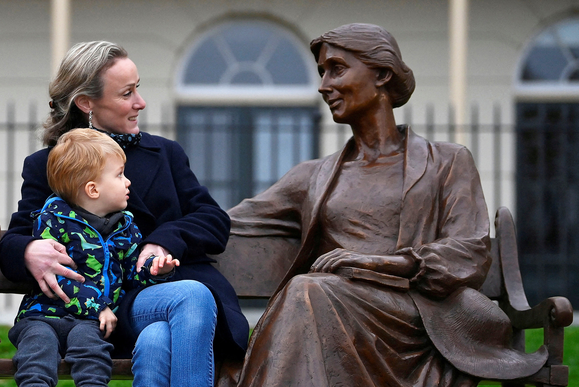 a woman and her young child sit on a bench looking warmly at a statue of a woman who is also sitting on the bench