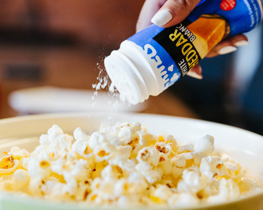 a person shaking white cheddar seasoning on to a bowl of popcorn