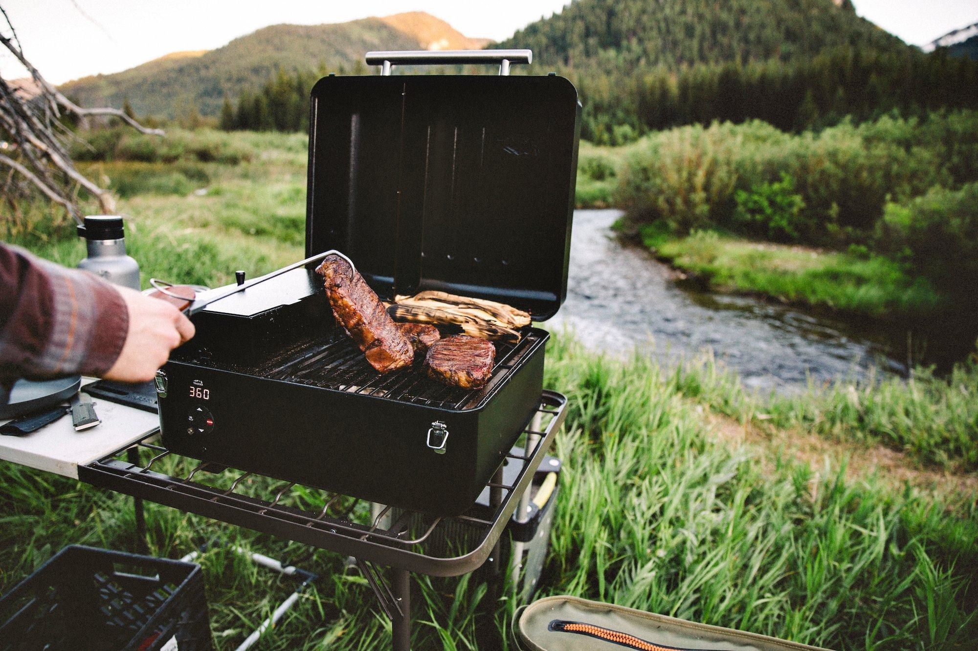 the portable grill set up next to a river and cooking steaks