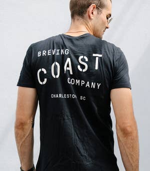 back of a model in a black t-shirt that reads "Brewing Coast Company - Charleston SC"